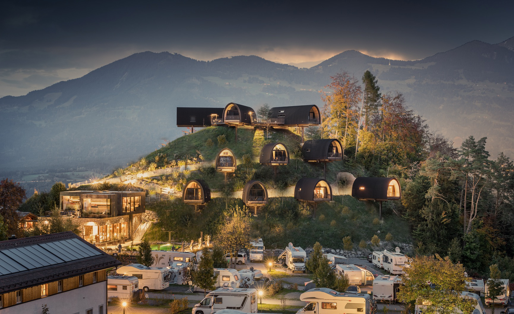 Luxuriöse Glamping-Lodges auf einem bewaldeten Hügel oberhalb eines modernen Campingplatzes bei Abenddämmerung, mit stimmungsvoller Beleuchtung und Bergen im Hintergrund.